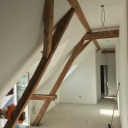 Poddasze w trakcie wykończenia: widoczne drewniane belki konstrukcyjne, świeżo pomalowane ściany na biało, surowa wylewka na podłodze, wystające przewody elektryczne i pozostawiona miotła.
