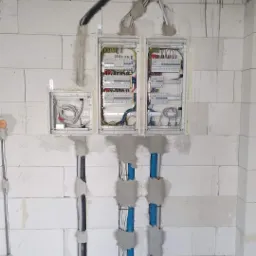Rozdzielnia elektryczna w trakcie montażu w surowym pomieszczeniu z białych bloczków, widoczne kable w peszlach i w otwartych puszkach, częściowo zamurowane.