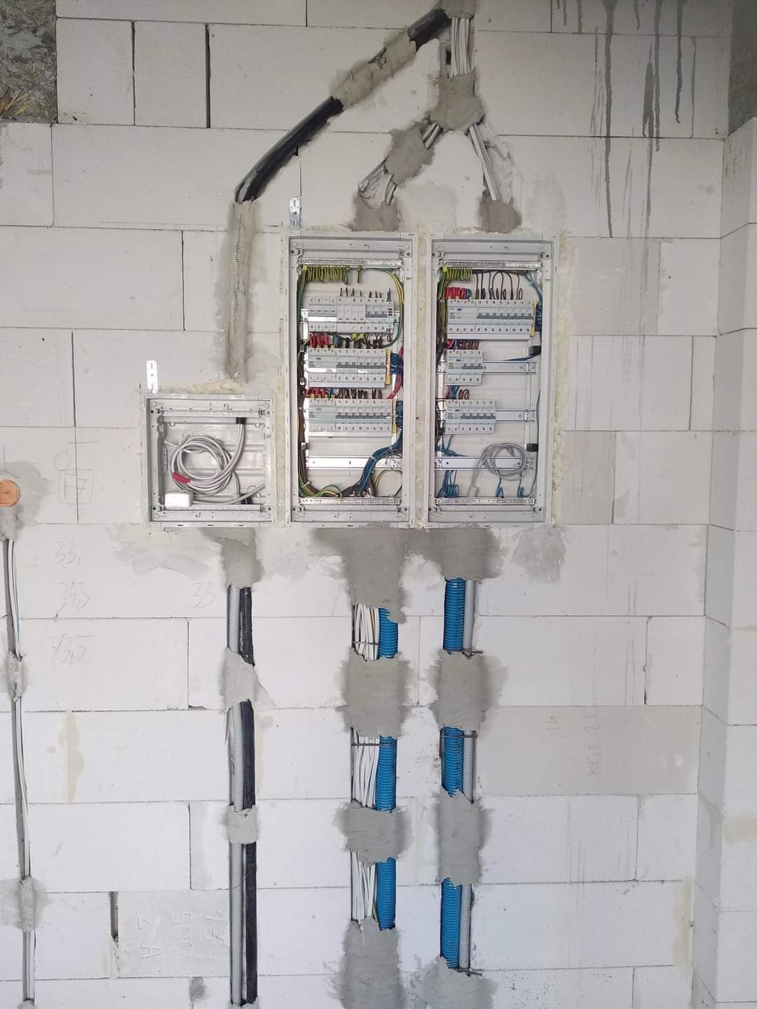 Rozdzielnia elektryczna w trakcie montażu w surowym pomieszczeniu z białych bloczków, widoczne kable w peszlach i w otwartych puszkach, częściowo zamurowane.