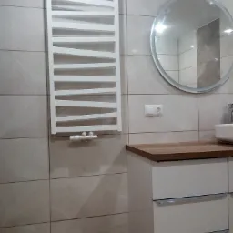 Nowoczesna łazienka z grzejnikiem drabinkowym, okrągłym lustrem i szafką pod umywalką z drewnianym blatem, wykończona płytkami imitującymi kamień.