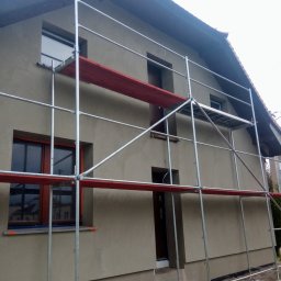 MKT-BUD Mariusz Burkowicz - Dom jednorodzinny w trakcie wykonywania elewacji, widoczne rusztowanie i świeżo nałożony tynk w kolorze beżowym, brązowe ramy okienne.