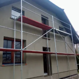 Dom jednorodzinny w trakcie wykonywania elewacji, widoczne rusztowanie i świeżo nałożony tynk w kolorze beżowym, brązowe ramy okienne.