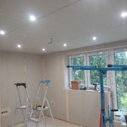 MKT-BUD Mariusz Burkowicz - Pokój w trakcie prac wykończeniowych: ściany i sufit pokryte płytami gipsowo-kartonowymi, widoczne dwie drabiny, podnośnik do płyt oraz narzędzia na parapecie okna.