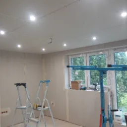 Pokój w trakcie prac wykończeniowych: ściany i sufit pokryte płytami gipsowo-kartonowymi, widoczne dwie drabiny, podnośnik do płyt oraz narzędzia na parapecie okna.