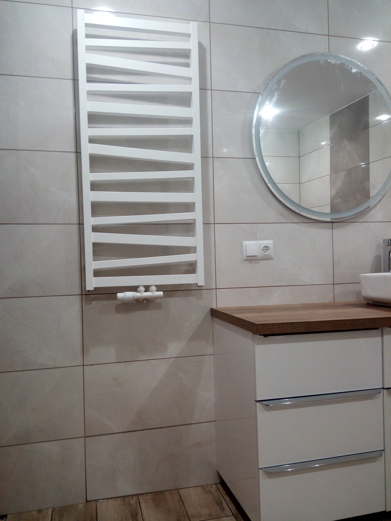 Nowoczesna łazienka z grzejnikiem drabinkowym, okrągłym lustrem i szafką pod umywalką z drewnianym blatem, wykończona płytkami imitującymi kamień.
