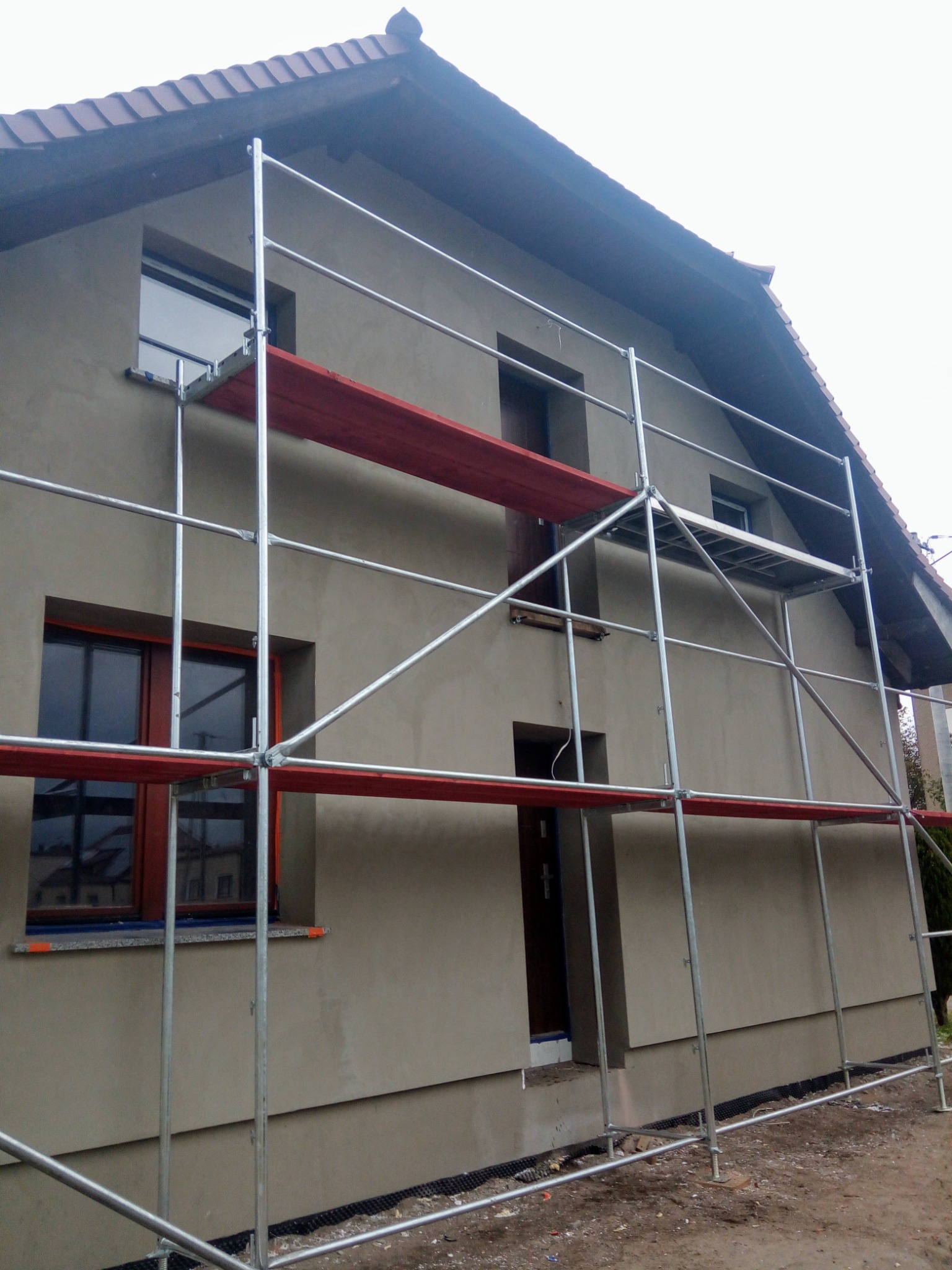 Dom jednorodzinny w trakcie wykonywania elewacji, widoczne rusztowanie i świeżo nałożony tynk w kolorze beżowym, brązowe ramy okienne.