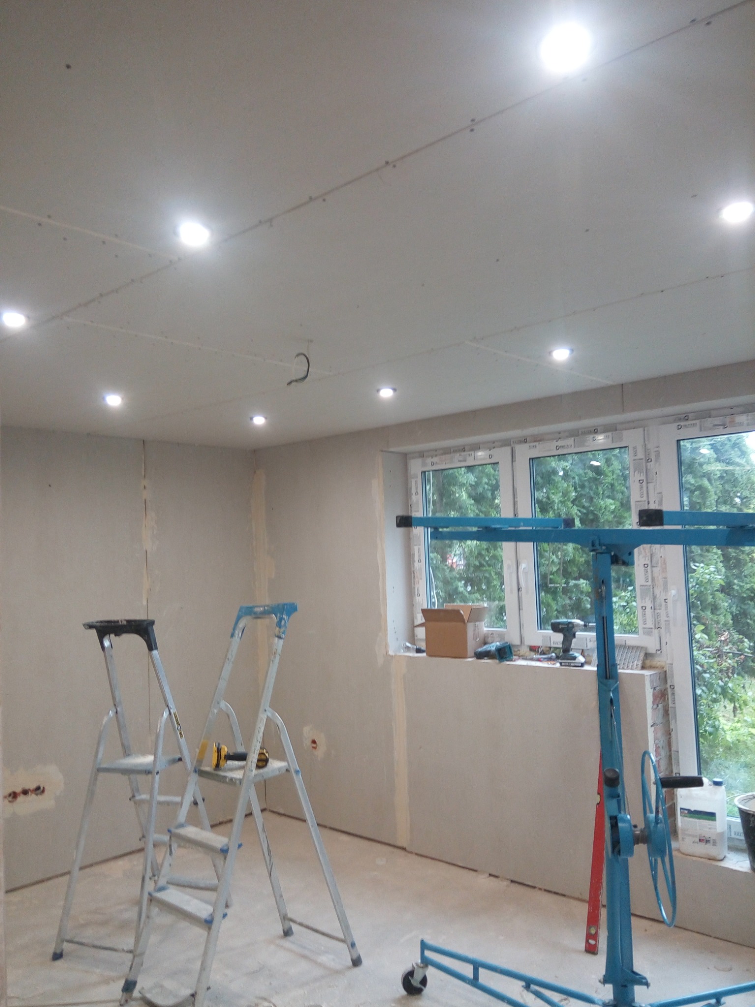 Pokój w trakcie prac wykończeniowych: ściany i sufit pokryte płytami gipsowo-kartonowymi, widoczne dwie drabiny, podnośnik do płyt oraz narzędzia na parapecie okna.