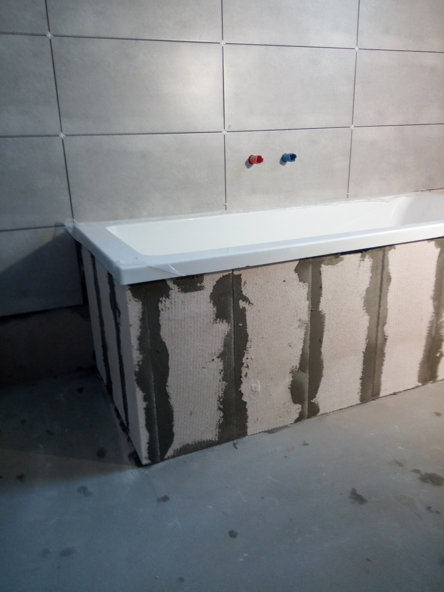 Wanna obudowana bloczkami z betonu komórkowego, z widocznymi pionowymi pasami zaprawy, przy ścianie wyłożonej szarymi płytkami z krzyżykami dystansowymi i wystającymi rurami instalacji wodnej.