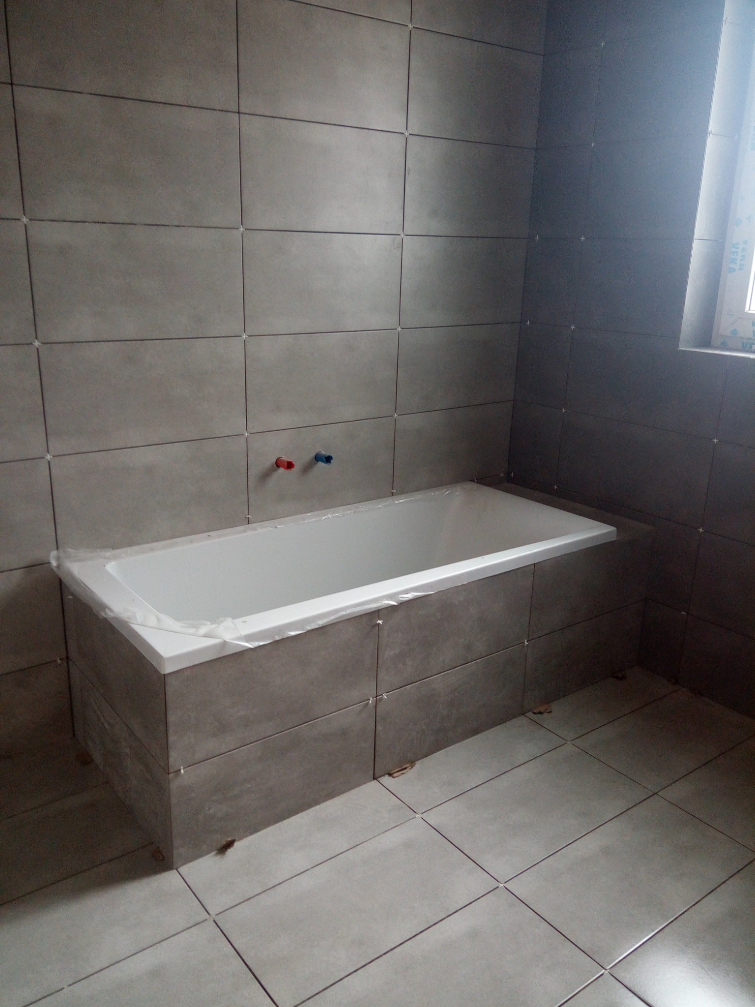 Szare, prostokątne płytki ścienne i podłogowe w łazience, biała wanna zabezpieczona folią, widoczne krzyżyki dystansowe, wystające rury od baterii.