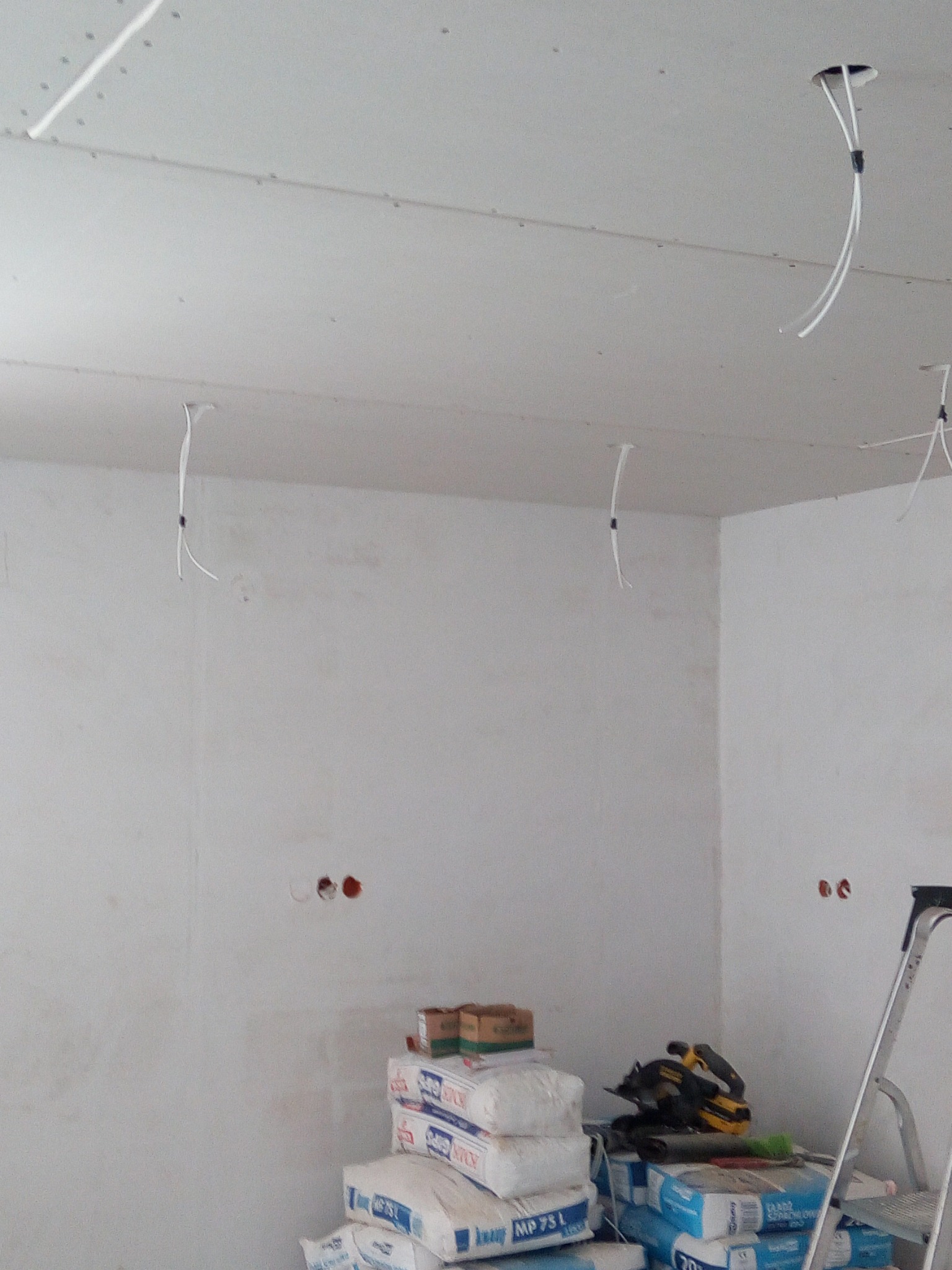 Przygotowania do wykończenia wnętrza w Klonowej: ściany po gładzi, widoczne kable wystające z sufitu, worki z zaprawą i drabina.