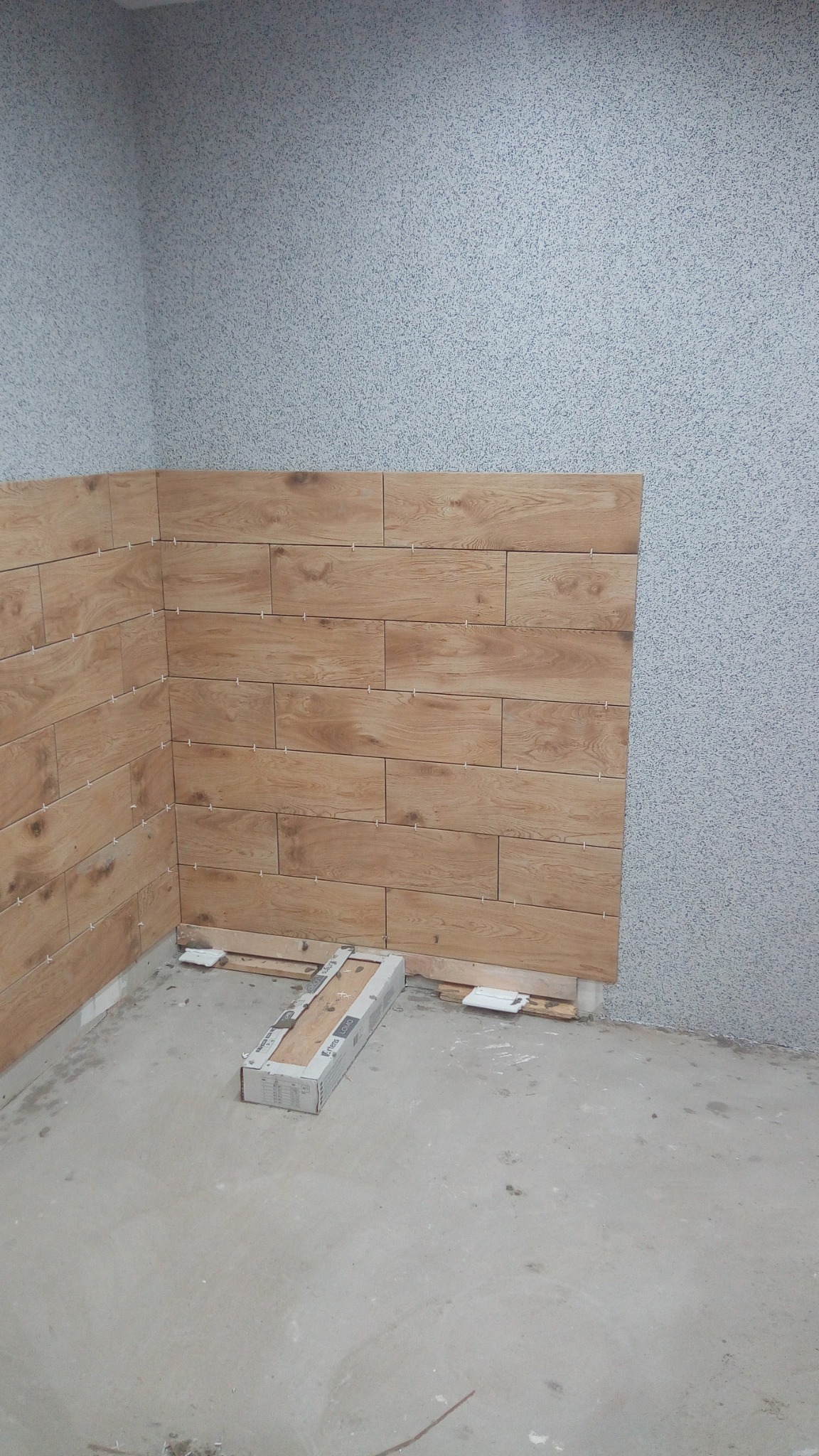 Układanie płytek drewnopodobnych na ścianie w rogu pomieszczenia, widoczna paczka płytek i krzyżyki dystansowe.