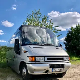 Srebrny bus turystyczny z polską tablicą rejestracyjną DDZ 12891, zaparkowany na żwirowym podłożu, z widocznym odbiciem chmur na szybie i zielenią w tle.