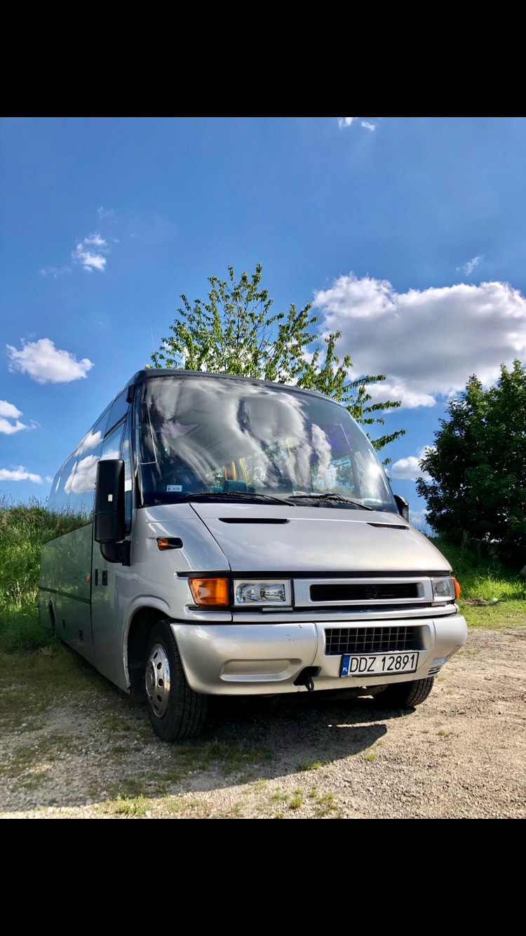 Srebrny bus turystyczny z polską tablicą rejestracyjną DDZ 12891, zaparkowany na żwirowym podłożu, z widocznym odbiciem chmur na szybie i zielenią w tle.