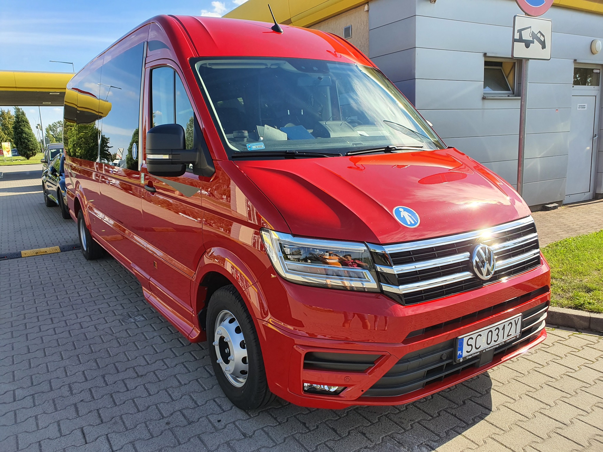 Czerwony Volkswagen Crafter z symbolem osoby na wózku inwalidzkim, zaparkowany na kostce brukowej przed budynkiem stacji obsługi.