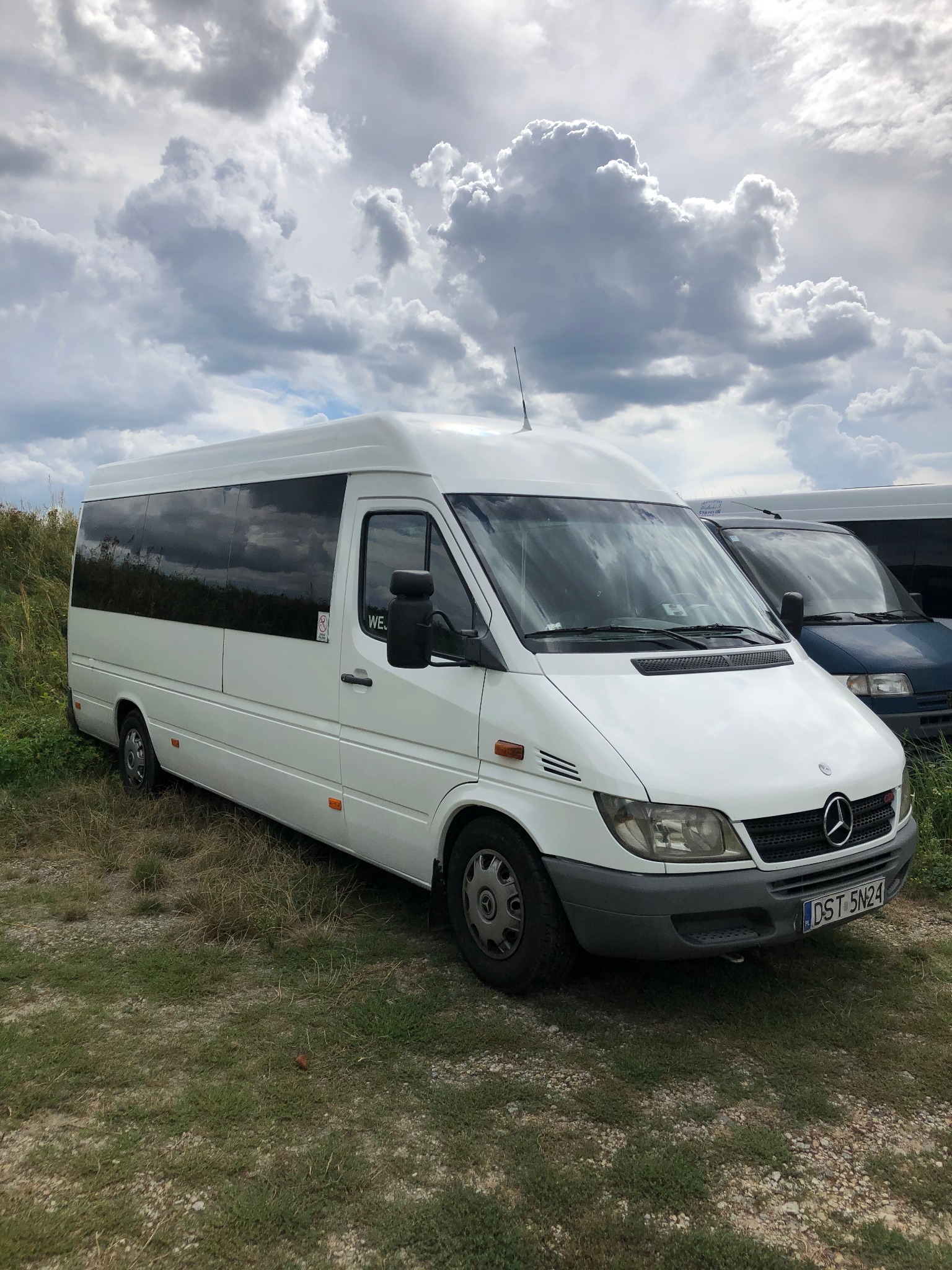 Biały bus Mercedes Sprinter z przyciemnianymi szybami, zaparkowany na trawiastym terenie pod pochmurnym niebem. Widoczna tablica rejestracyjna i fragment drugiego pojazdu z tyłu.