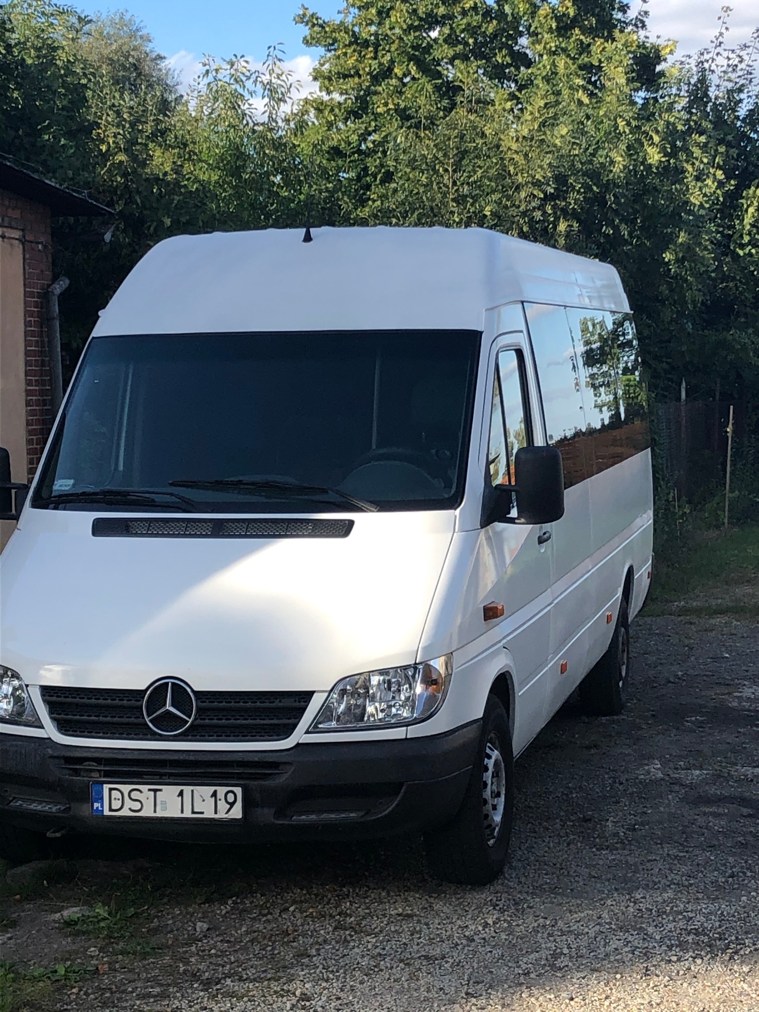 Biały bus Mercedes Sprinter z polskimi tablicami rejestracyjnymi DST 1L19, zaparkowany na żwirowym podłożu, z widocznym fragmentem ceglanego budynku i zieleni w tle.