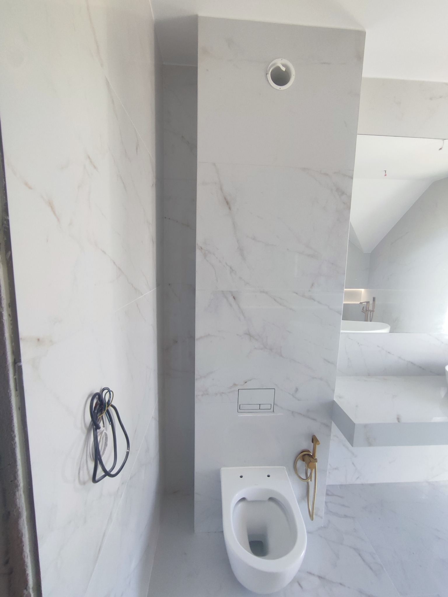 Nowoczesna łazienka z białymi płytkami imitującymi marmur, wisząca toaleta i złota armatura. Widoczny fragment wanny w tle.