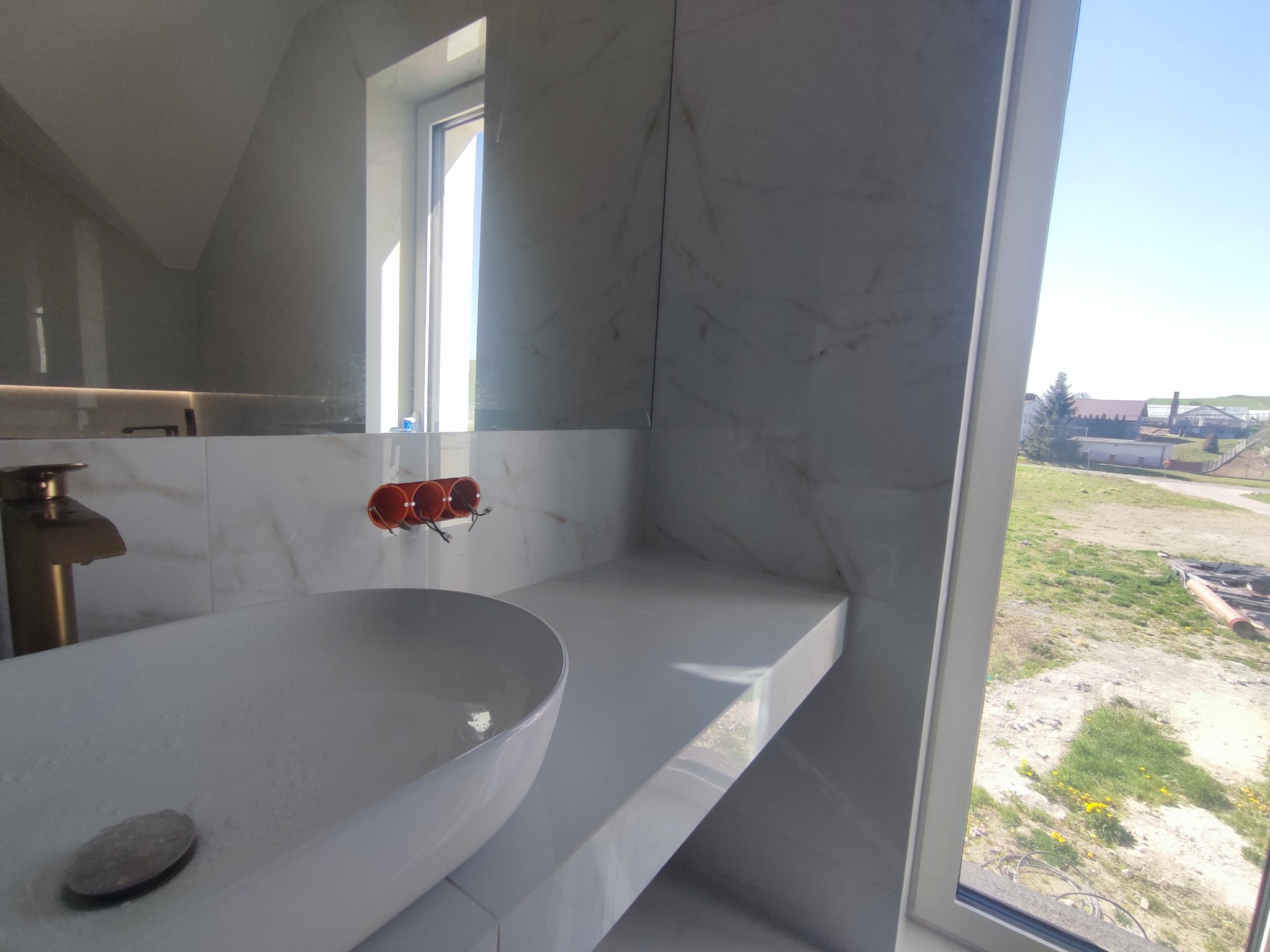 Biała, owalna umywalka nablatowa z baterią w kolorze złota na blacie z płytek imitujących marmur. W tle okno z widokiem na zewnątrz i instalacja wodna.