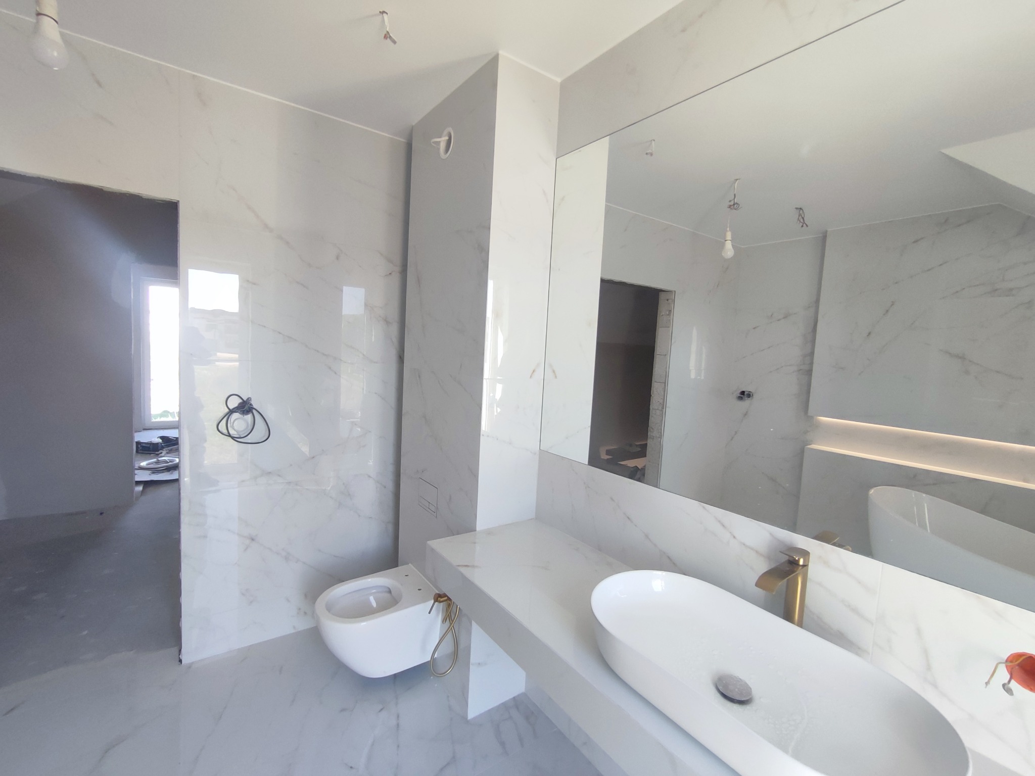 Nowoczesna łazienka z białymi płytkami imitującymi marmur, wisząca toaleta i umywalka nablatowa z złotą baterią, widok na niedokończone pomieszczenie.