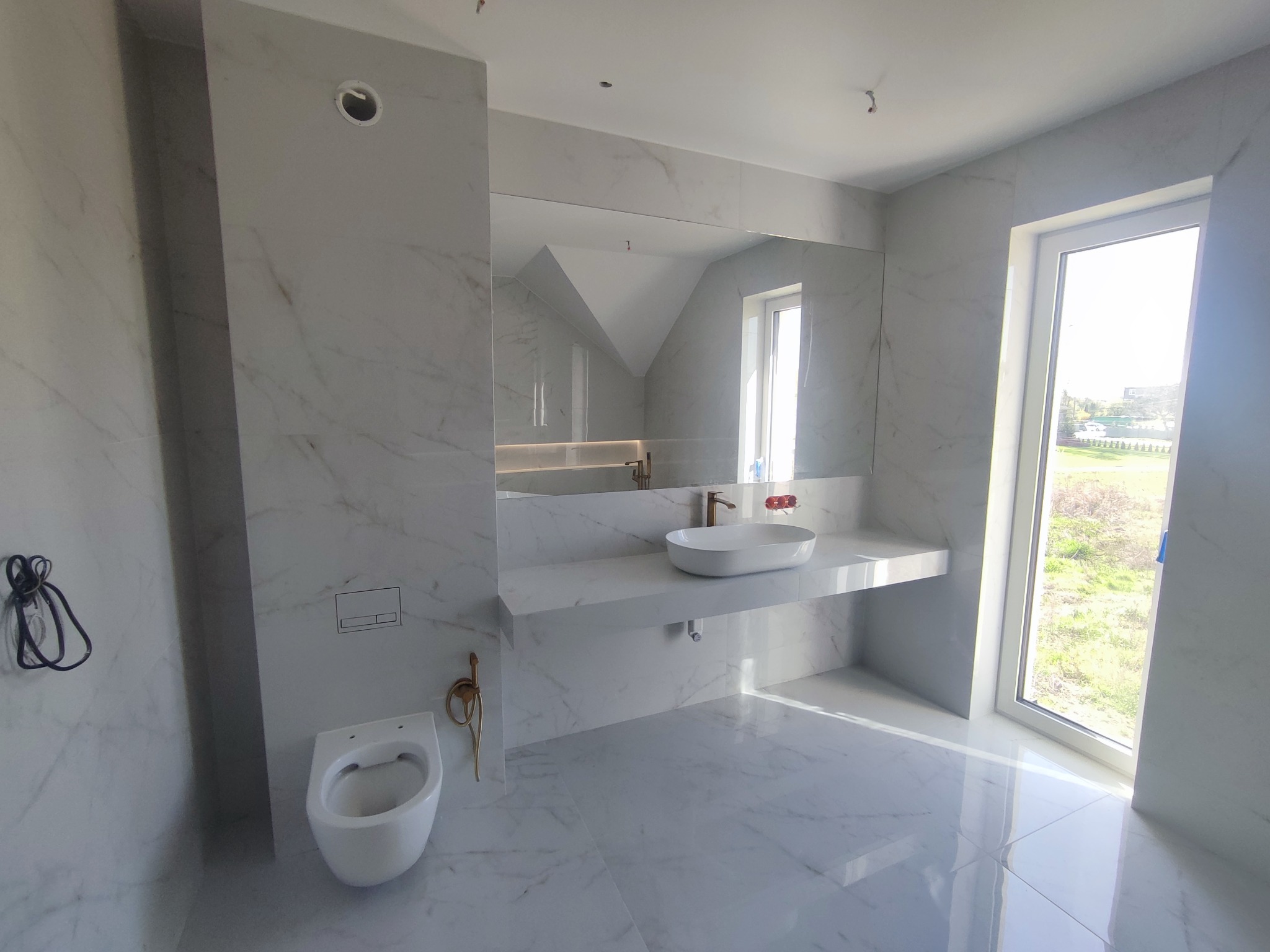 Jasna łazienka z białymi płytkami imitującymi marmur, wisząca miska WC i umywalka na blacie. Widoczne okno z widokiem na zieleń. Złote elementy armatury.