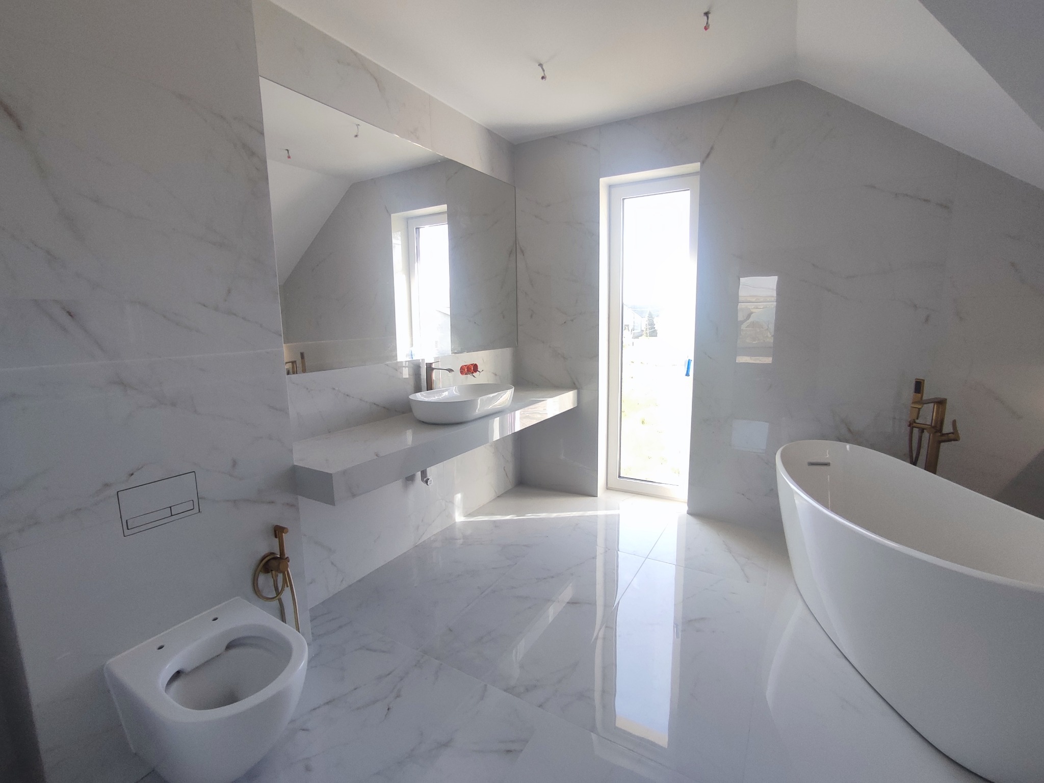 Elegancka łazienka na poddaszu w bieli: marmurowe płytki, wolnostojąca wanna, umywalka nablatowa i złota armatura. Jasne wnętrze z oknem.