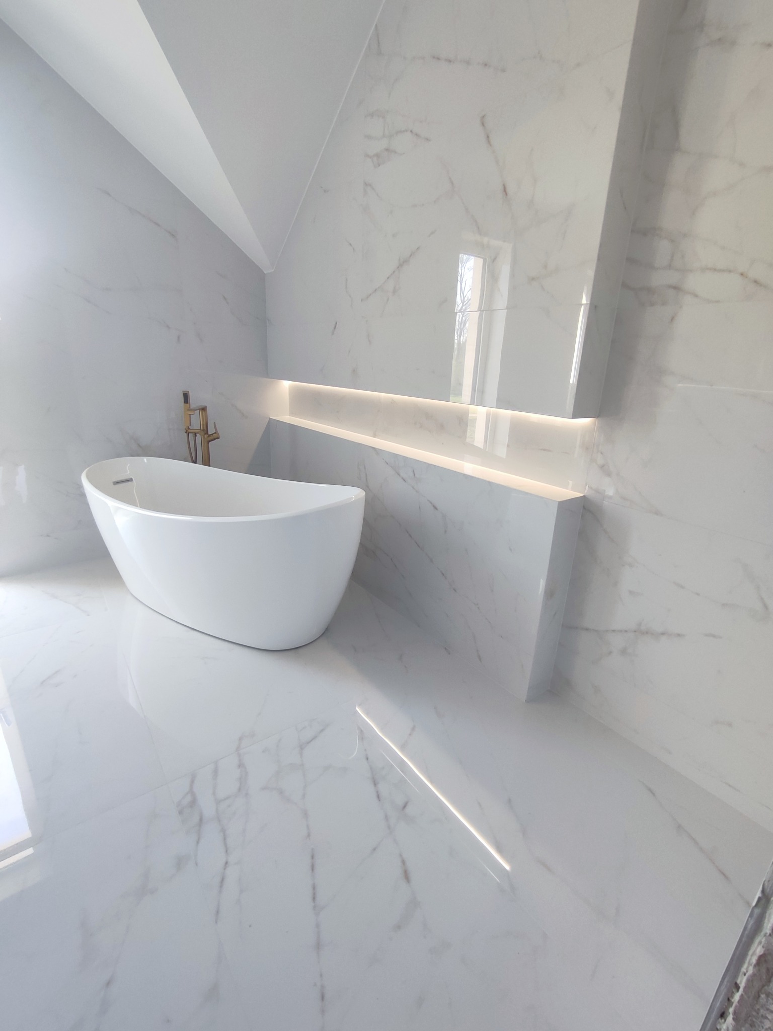 Nowoczesna łazienka z wolnostojącą wanną i marmurowymi płytkami. Podświetlana półka w ścianie dodaje elegancji. Minimalistyczny design, jasne wnętrze.