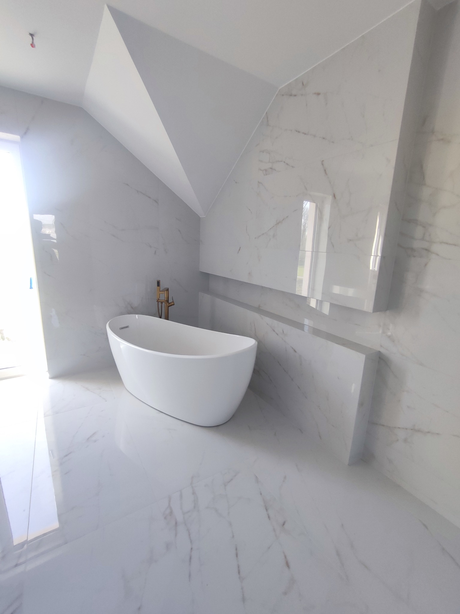 Biała, wolnostojąca wanna z złotą armaturą w nowoczesnej łazience na poddaszu. Ściany i podłoga wyłożone błyszczącymi, marmurowymi płytami.