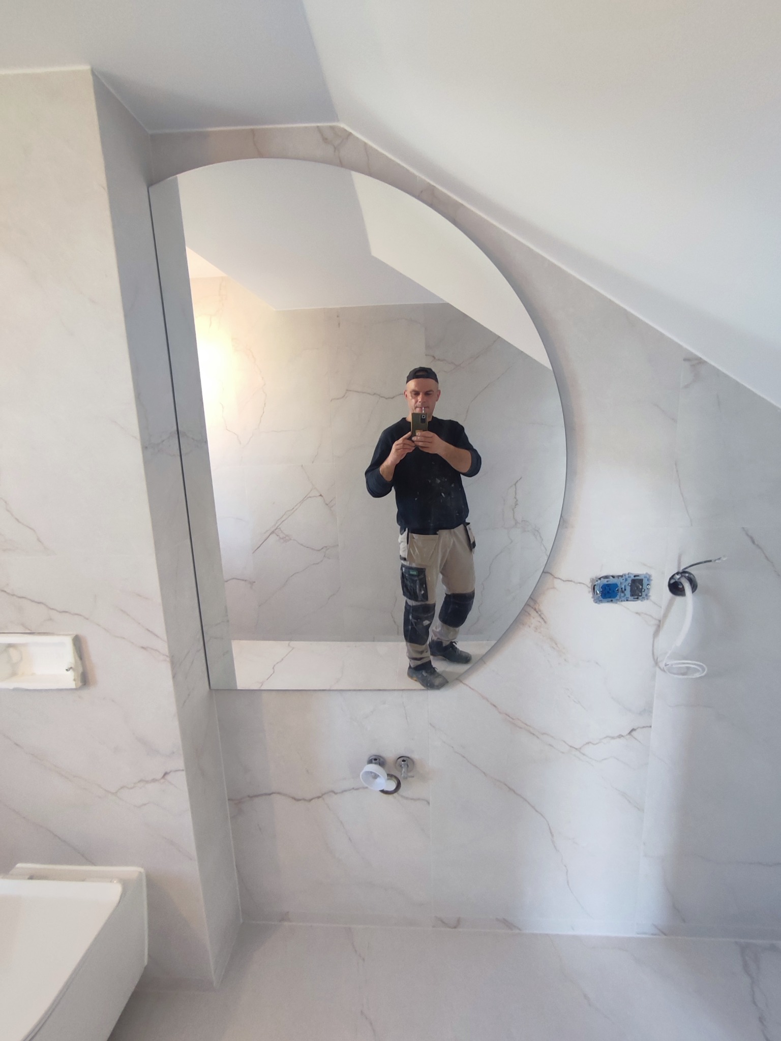 Wykończona łazienka z marmurowymi płytkami i owalnym lustrem, w którym odbija się glazurnik robiący zdjęcie telefonem. Widoczne gniazdka elektryczne i instalacja wodna.