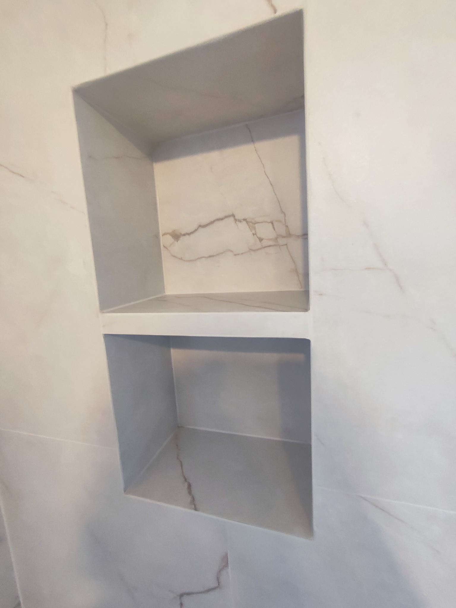 Wnęka w łazience wyłożona płytkami imitującymi marmur, z widocznymi żyłkami. Dwie półki tworzą praktyczne miejsce do przechowywania.