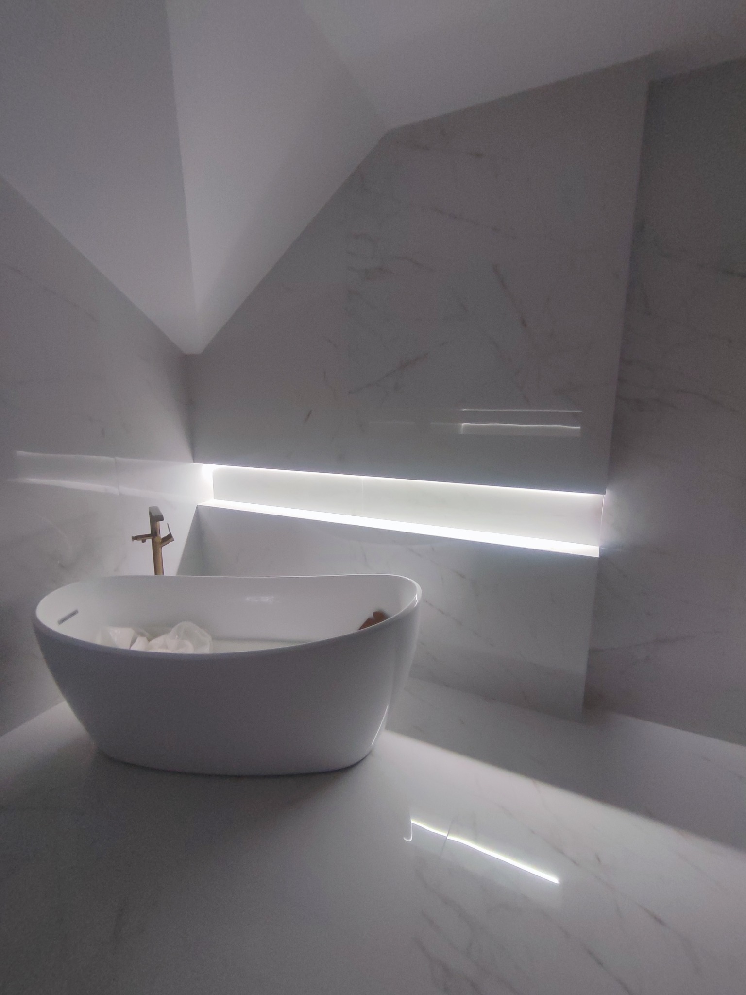 Elegancka, biała wanna wolnostojąca z złotą armaturą w nowoczesnej łazience z marmurowymi płytkami i subtelnym oświetleniem LED w zabudowie.