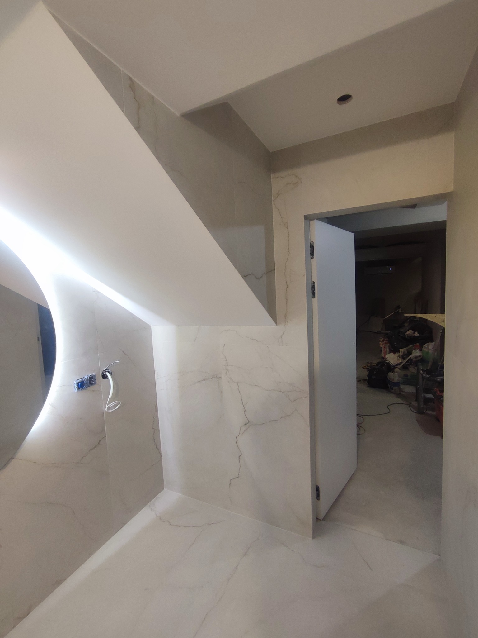 Wykończona łazienka z płytkami imitującymi marmur, oświetlonym lustrem i drzwiami. Widoczny fragment sufitu podwieszanego.