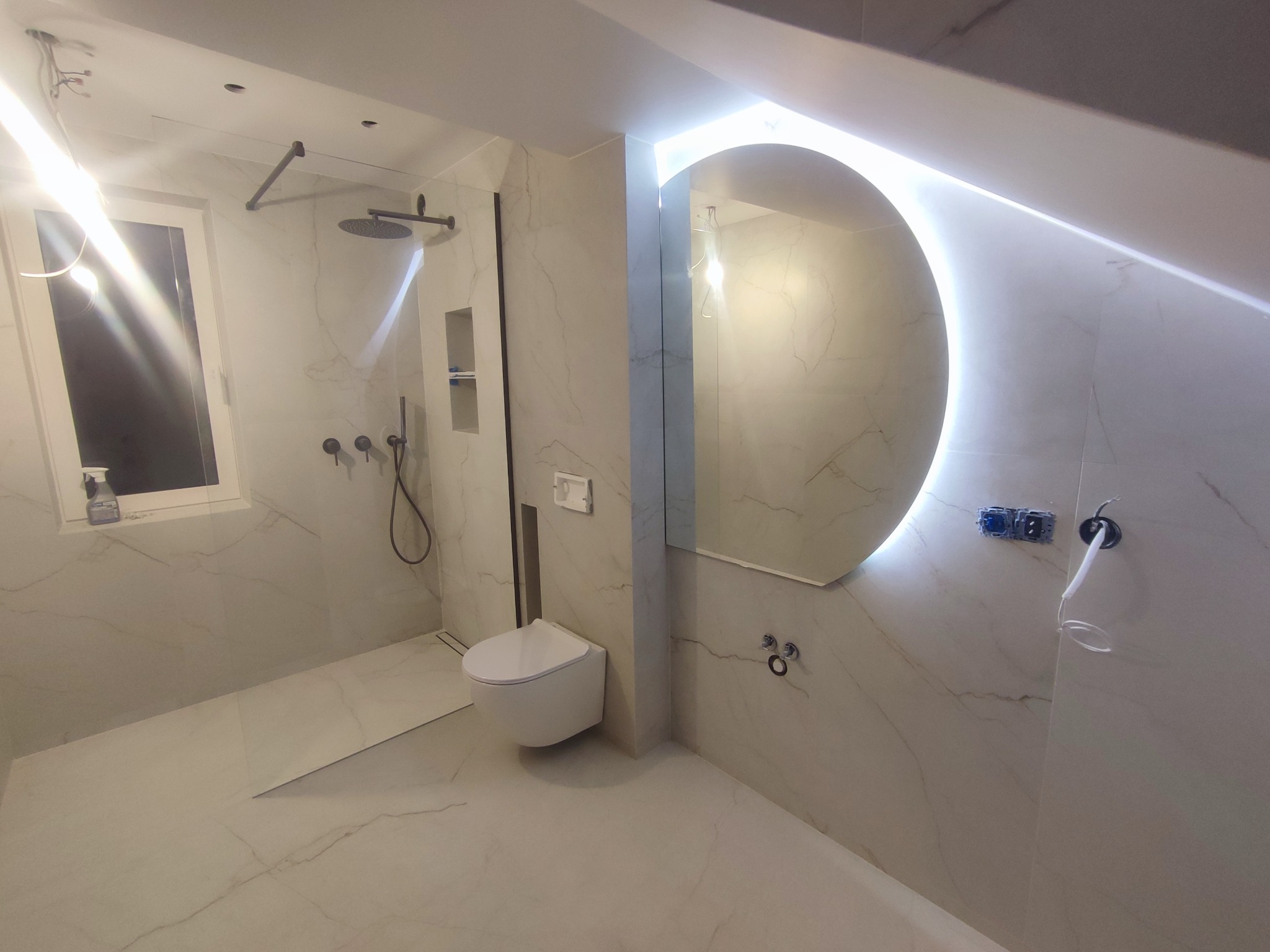 Wykończona łazienka na poddaszu z kabiną typu walk-in, podwieszaną toaletą i okrągłym lustrem z podświetleniem LED. Białe płytki imitujące marmur na ścianach i podłodze.