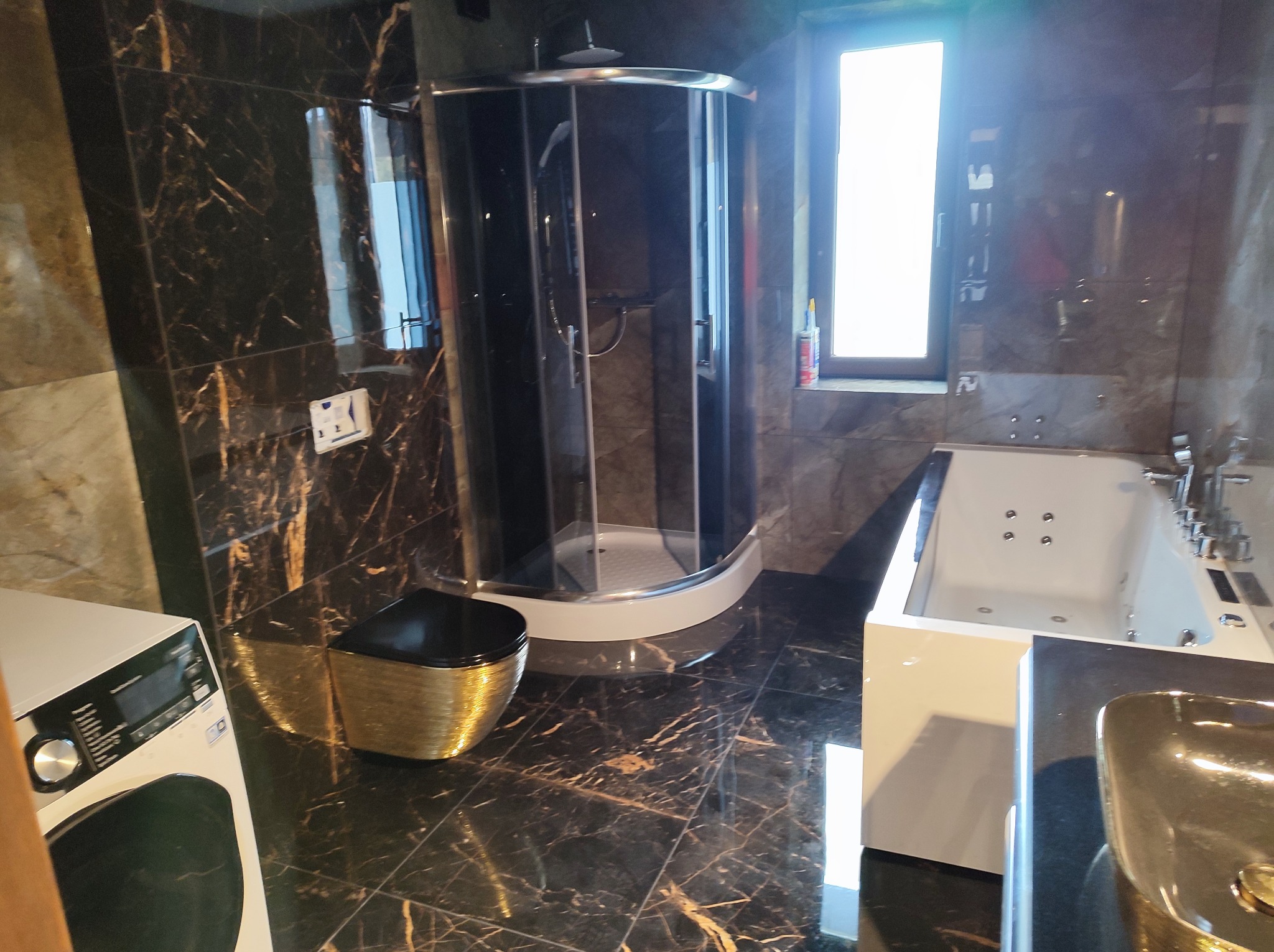 Elegancka łazienka z czarnymi płytkami imitującymi marmur, kabiną prysznicową, wanną z hydromasażem i złotą umywalką. Widoczne okno z jasnym światłem.