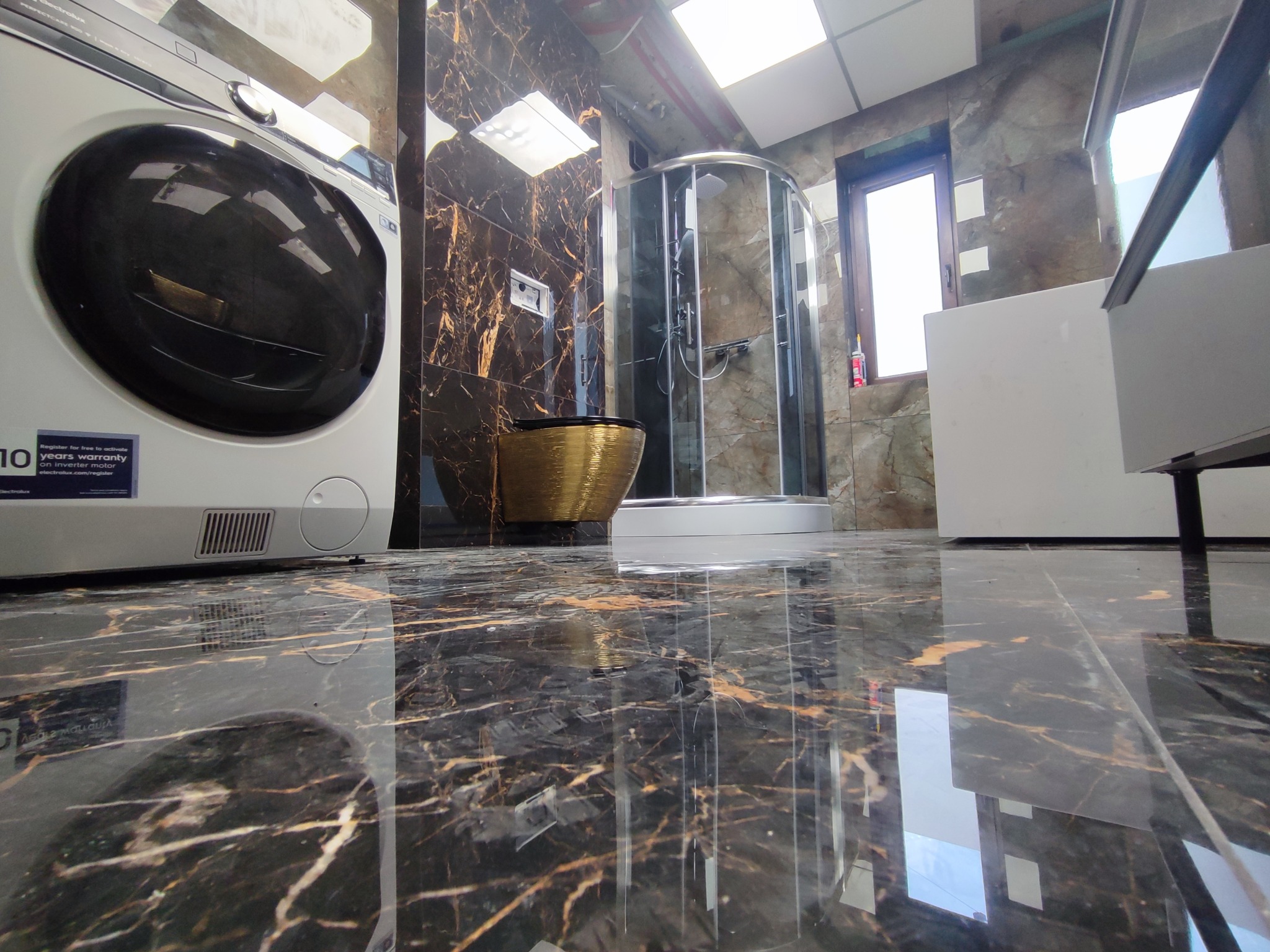 Elegancka łazienka z kabiną prysznicową, złotą miską WC i pralką. Uwagę przyciągają marmurowe płytki na ścianach i podłodze z połyskiem.