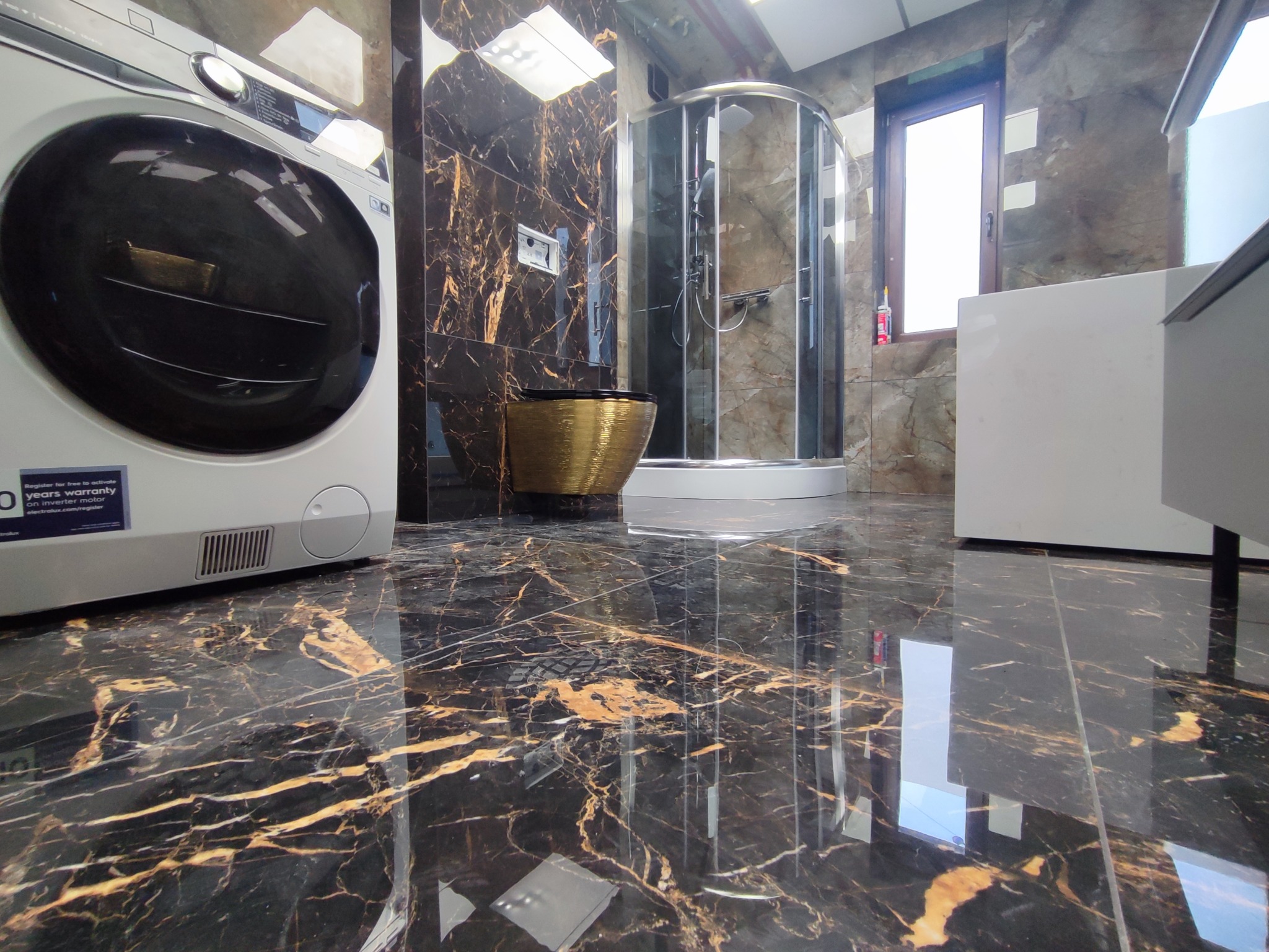 Luksusowa łazienka z kabiną prysznicową, złotą umywalką i pralką na tle marmurowych ścian i podłóg z wyraźnym połyskiem, okno z widoczną butelką silikonu.