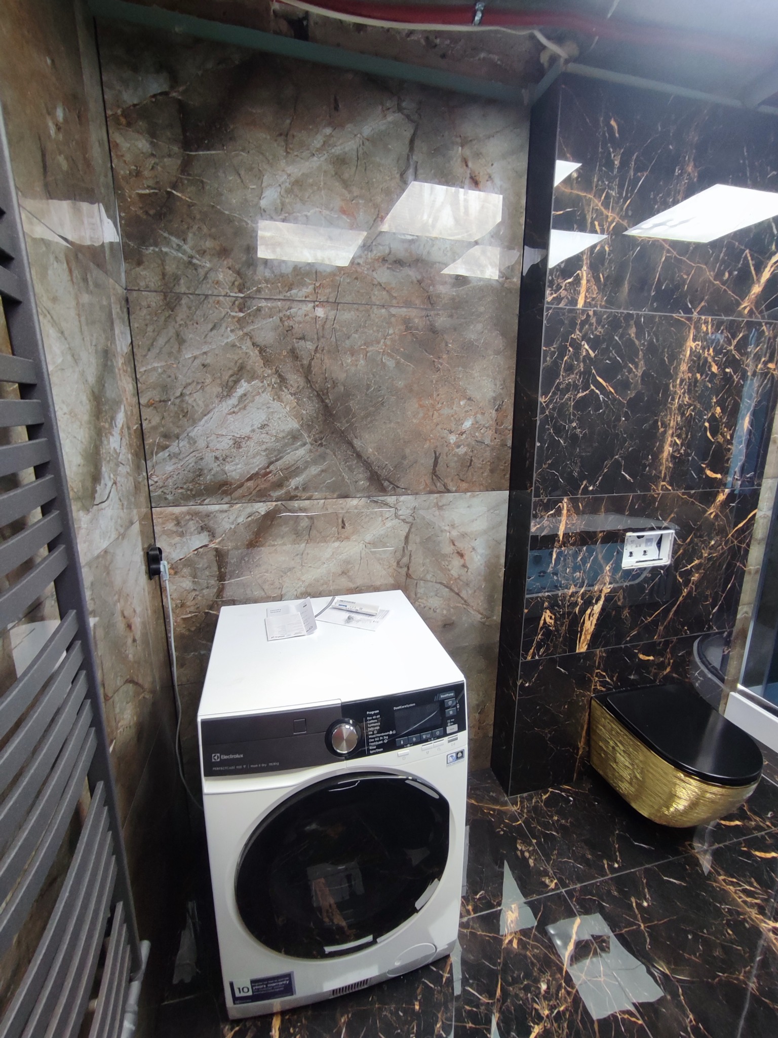 Nowoczesna łazienka z marmurowymi płytkami, białą pralką Electrolux i złotą toaletą. Grzejnik drabinkowy po lewej stronie. Eleganckie wykończenie.