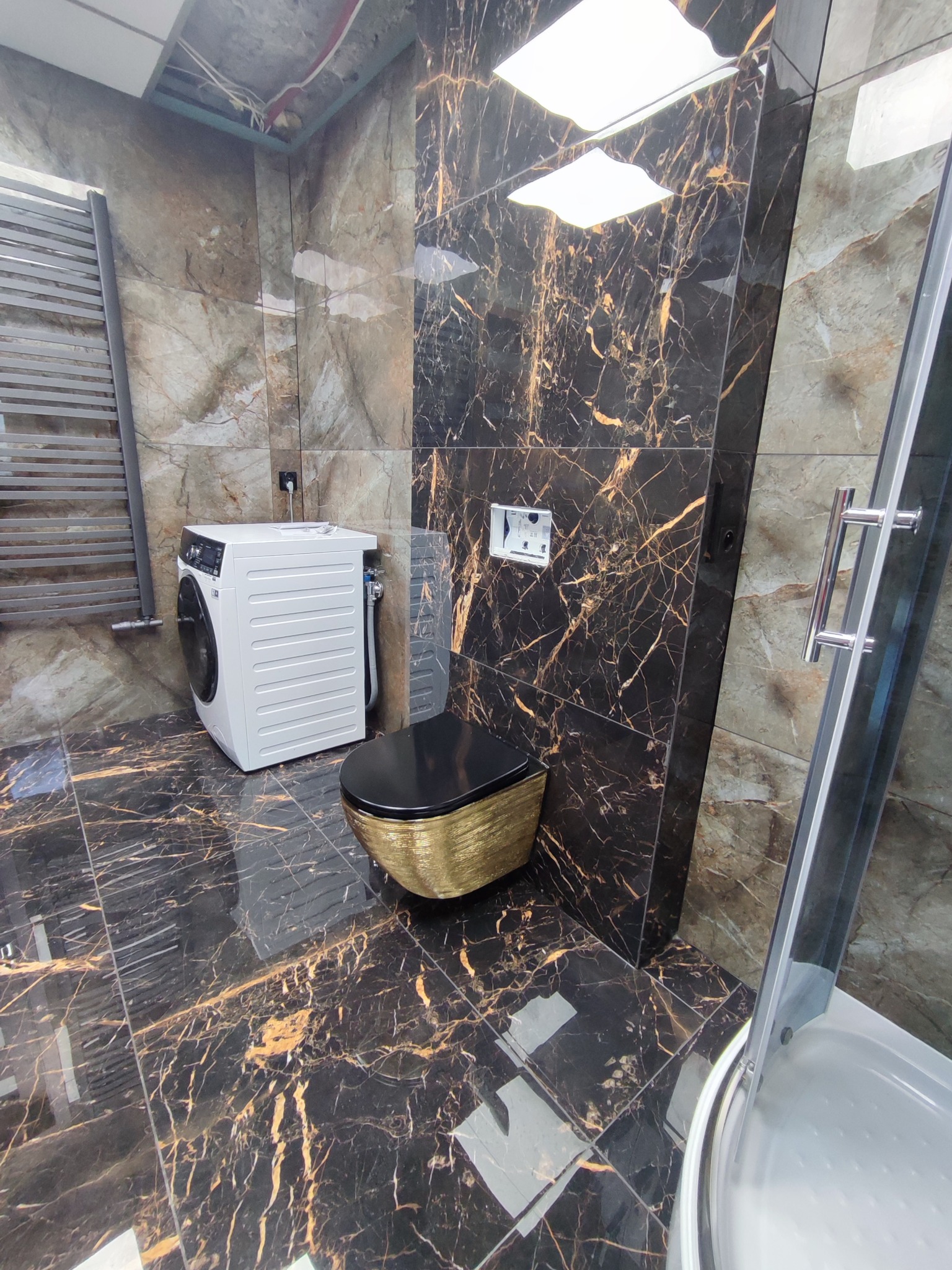 Elegancka łazienka z płytkami imitującymi marmur w odcieniach czerni i złota, wyposażona w pralkę, wiszącą toaletę i kabinę prysznicową.