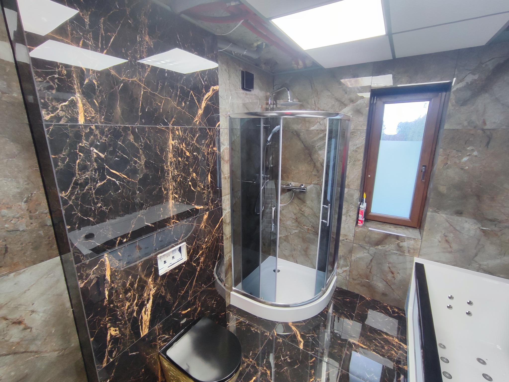 Elegancka łazienka z kabiną prysznicową, wykończona płytkami imitującymi marmur w odcieniach brązu i złota. Widoczna toaleta i wanna z hydromasażem.