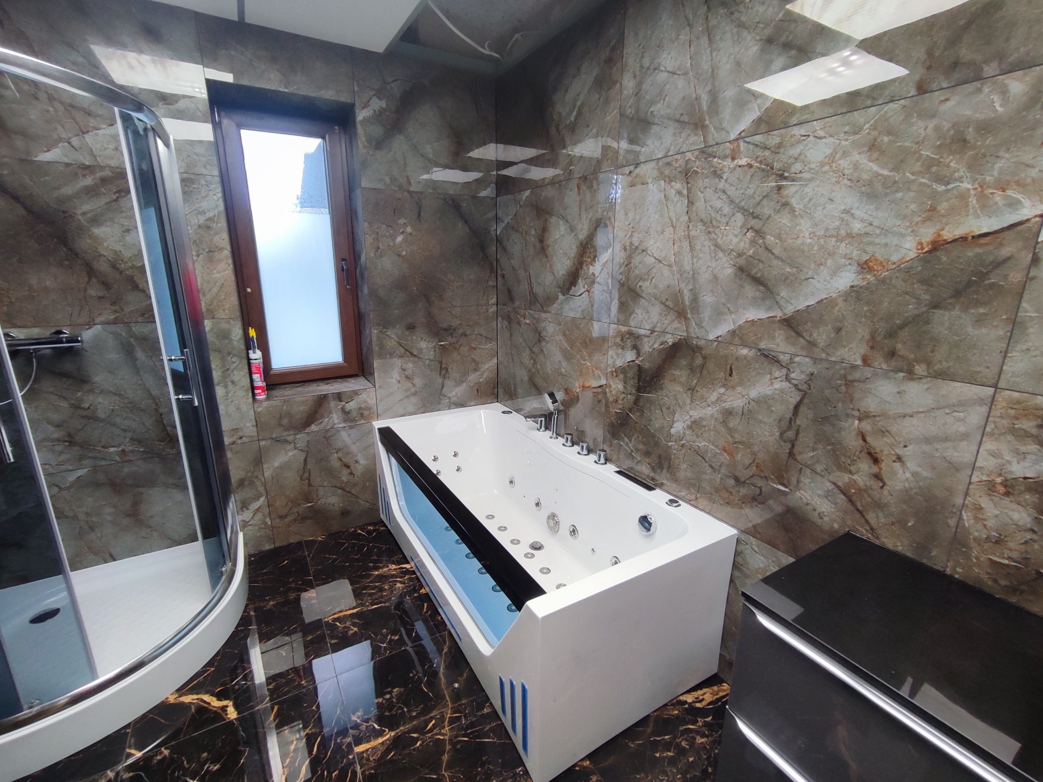 Luksusowa łazienka z kabiną prysznicową i wanną z hydromasażem, wykończona marmurowymi płytkami w odcieniach brązu i czerni, z ciemną, błyszczącą podłogą.