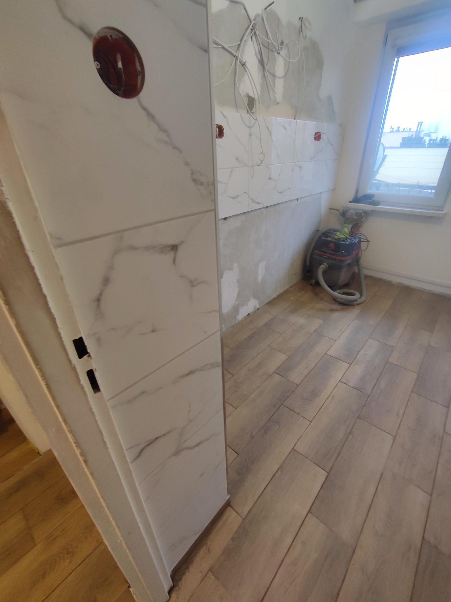 Wykończenie łazienki w Olkuszu: białe płytki imitujące marmur na ścianie, drewnopodobna podłoga, widoczne gniazdko elektryczne i odkurzacz.