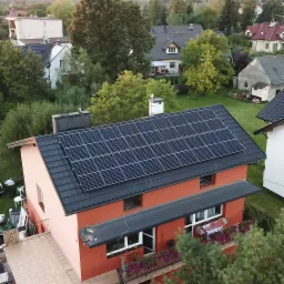 Widok z góry na dom z panelami słonecznymi na dachu, otoczony zielenią i innymi budynkami mieszkalnymi w tle.