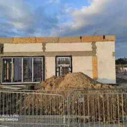 Wznoszenie ścian budynku z betonu komórkowego, widoczne otwory okienne i drzwiowe, zabezpieczone metalowymi ramami, na pierwszym planie hałda ziemi i metalowe barierki, w tle krajobraz z wieżą...