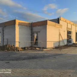 Budowa domu z białych bloczków, widoczne otwory okienne i drzwiowe, obrys dachu z desek, metalowe barierki zabezpieczające teren budowy, niebo z chmurami w tle.