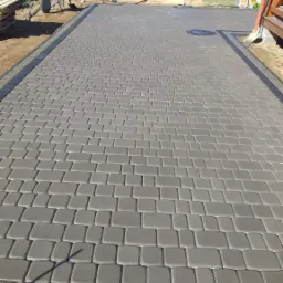 Szara, prostokątna kostka brukowa ułożona na posesji, widoczne palety z kostką i taczka w tle, jasny budynek gospodarczy.