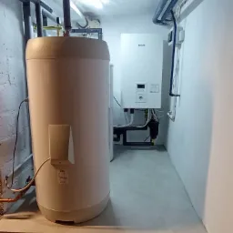 Bojler OSO oraz wiszący moduł Panasonic z podłączeniami w pomieszczeniu technicznym z szarymi ścianami i podłogą.