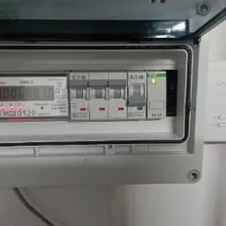 Otwarta skrzynka elektryczna z licznikiem zużycia energii i wyłącznikami nadprądowymi Eaton, widoczny także termostat Salus na ścianie.