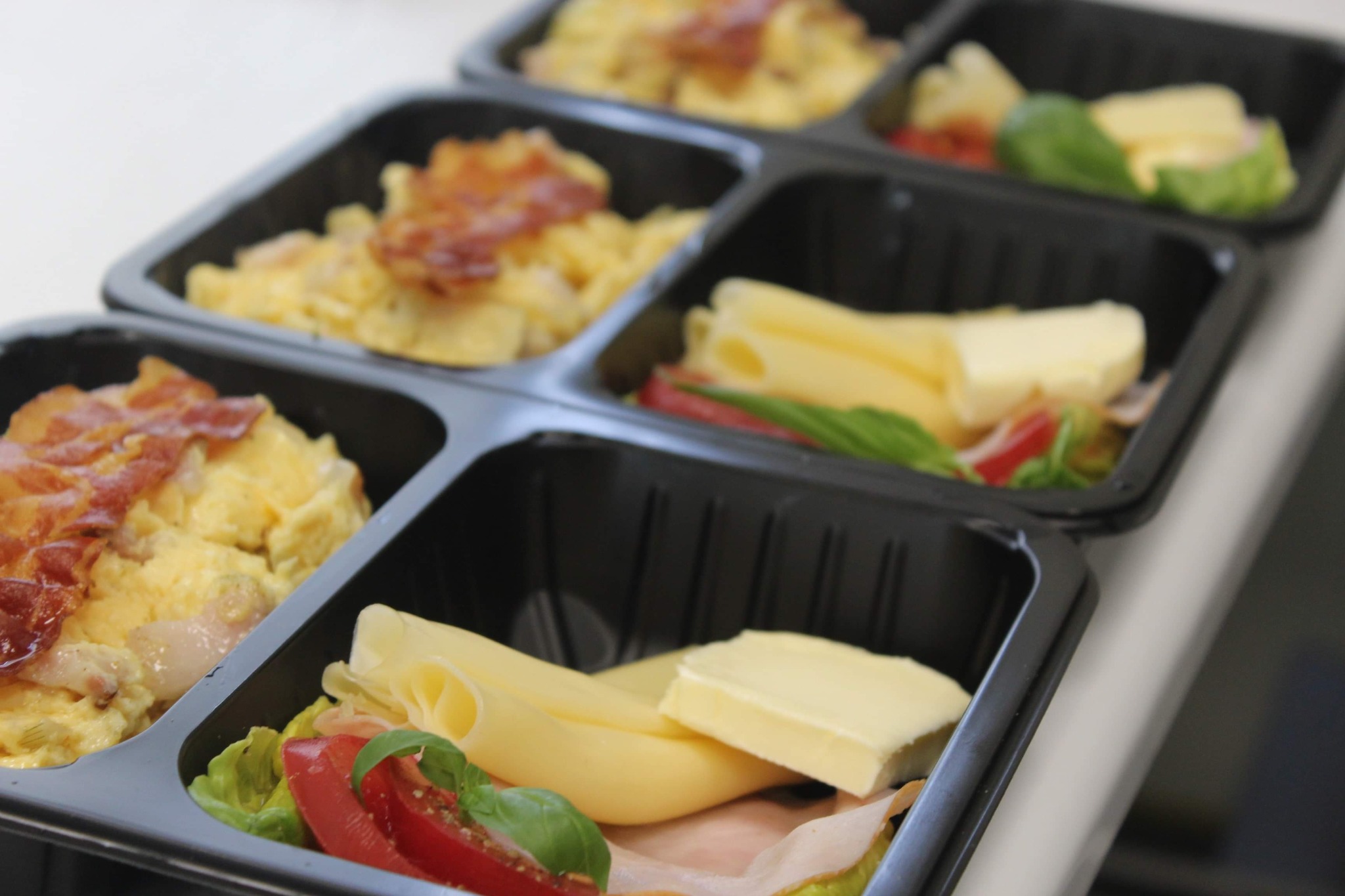 Czarne, plastikowe pojemniki z dwoma rodzajami posiłków: jajecznica z boczkiem oraz ser żółty i biały z szynką, pomidorem i bazylią. Catering dietetyczny w pudełkach.