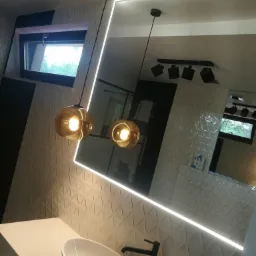 Nowoczesna łazienka z umywalką nablatową, lustrem podświetlonym LED i wiszącymi lampami ze szklanymi kloszami na tle ściany z trójkątną fakturą.