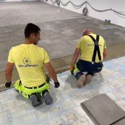 Dwóch glazurników w żółtych koszulkach z logo firmy układa szare płytki na podłodze, widoczne nierówności podłoża i kable pod sufitem.