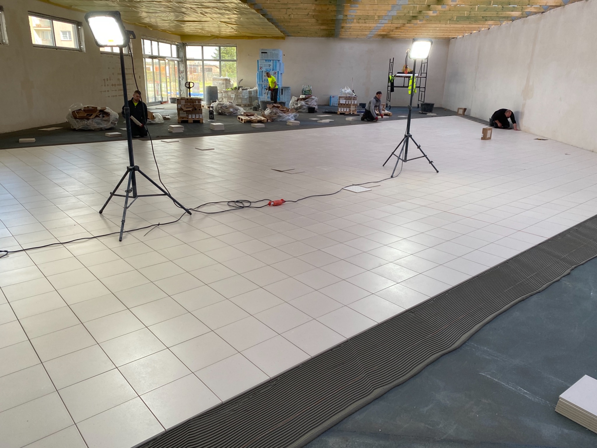 Montaż jasnych płytek podłogowych w dużym pomieszczeniu w Żarach, widoczne lampy budowlane, palety z materiałami i pracownicy w trakcie układania.